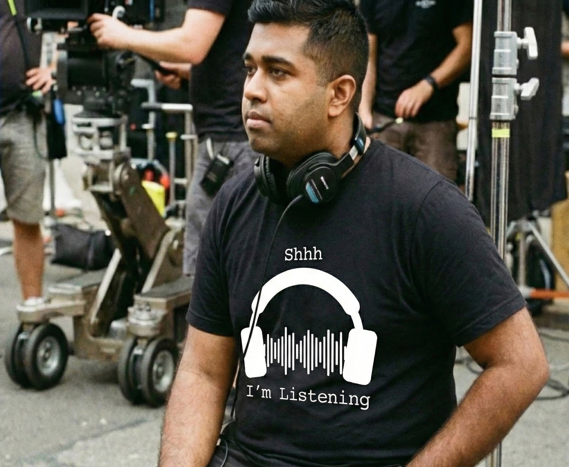 Person wearing a black t-shirt with headphones graphic and text, sitting on a set with filming equipment.